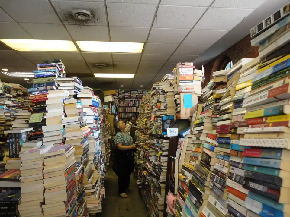 A woman stands in a narrow aisle of a cluttered bookstore, surrounded by towering, unorganized stacks of books that reach…