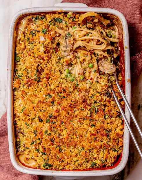 An overhead shot of a white ceramic baking dish filled with a creamy pasta casserole topped with golden-brown breadcrumbs…