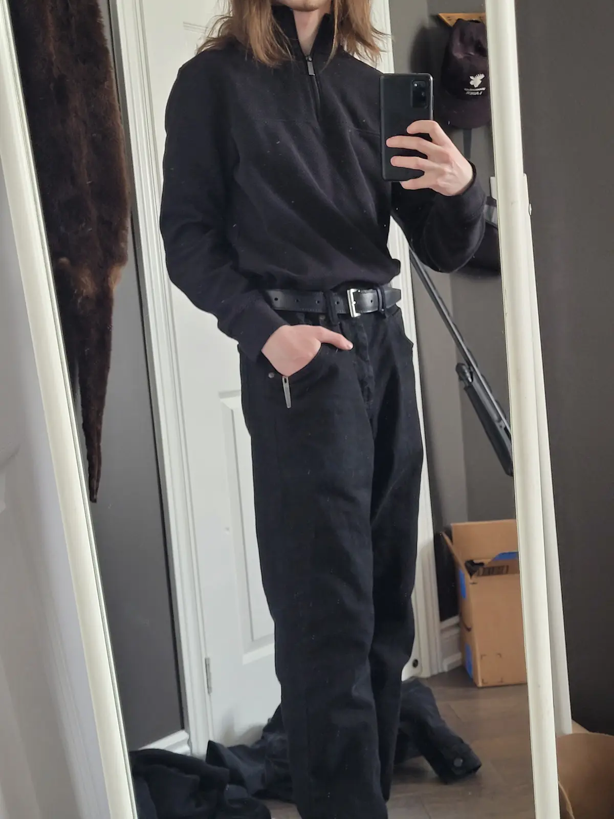 A person with long hair takes a mirror selfie wearing a black zip-neck sweater, black pants, and a black belt, with one hand…