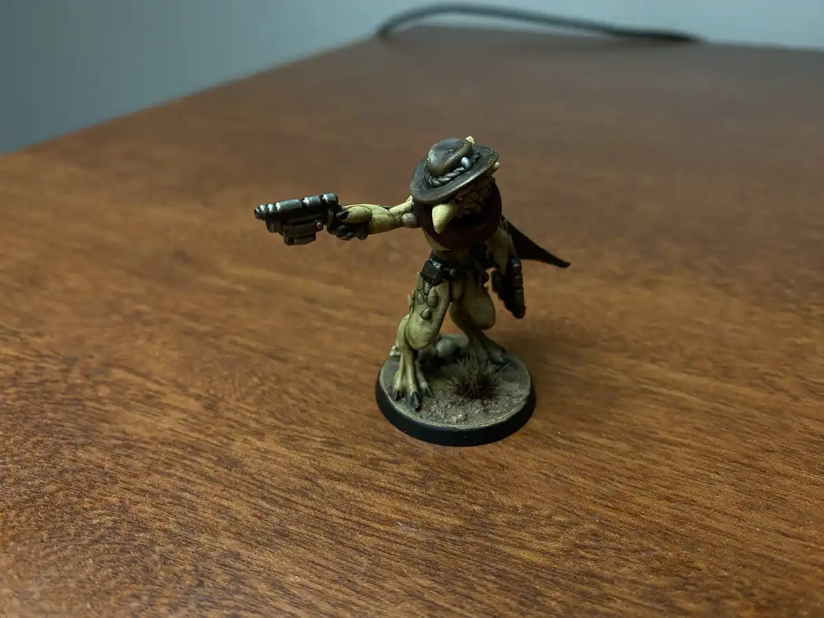 A painted miniature figurine of a bird-like humanoid wearing a hat and red scarf, holding a pistol and standing on a round…