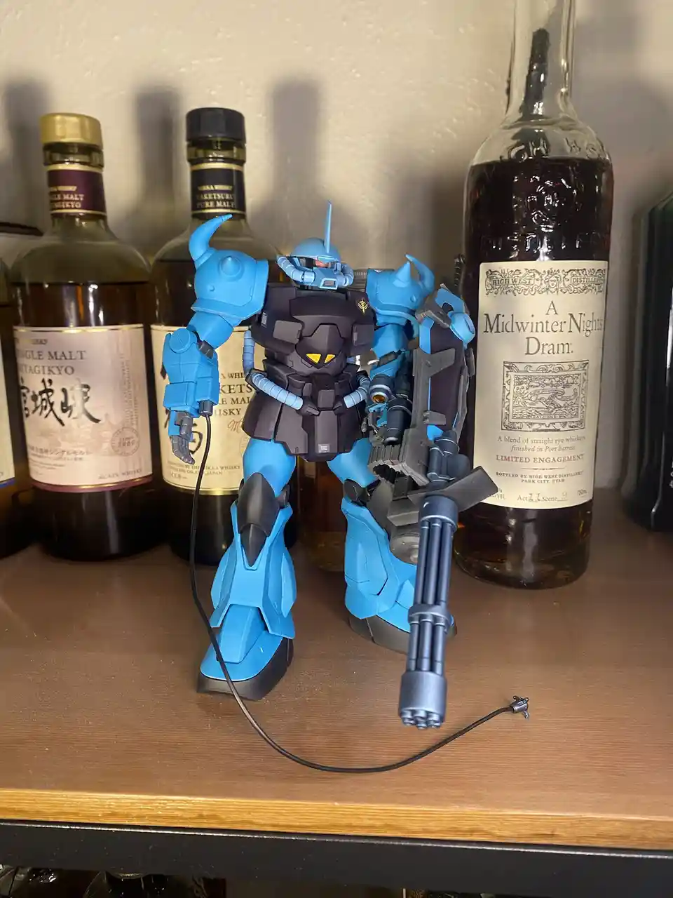 A blue and black robot model, resembling a Gundam, stands on a wooden shelf next to several whiskey bottles, including one…