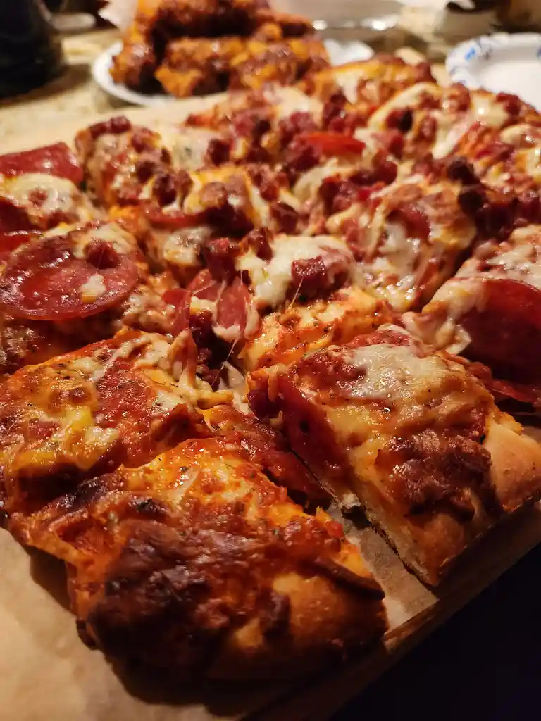 A close-up photo of a square-cut, cheesy pizza topped with pepperoni and other meats, served on parchment paper. The text…