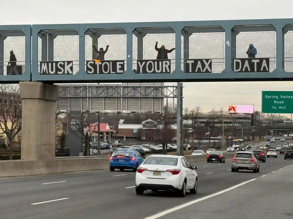 Several people stand on a pedestrian overpass above a busy highway, each positioned behind a section of a large sign that…