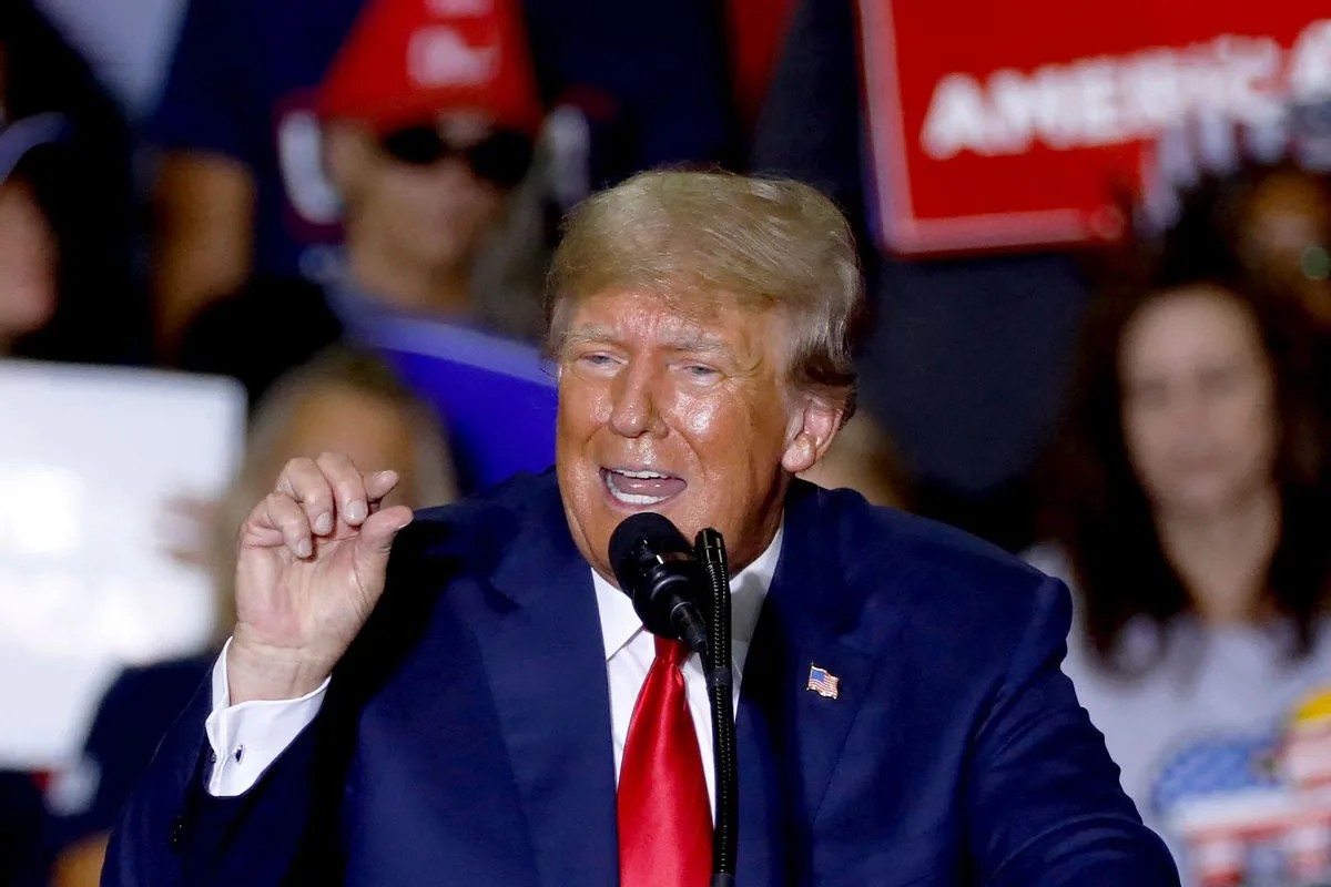 a photo of Donald Trump speaking at a rally, wearing a blue suit and red tie, with a microphone in front of him. Behind him…
