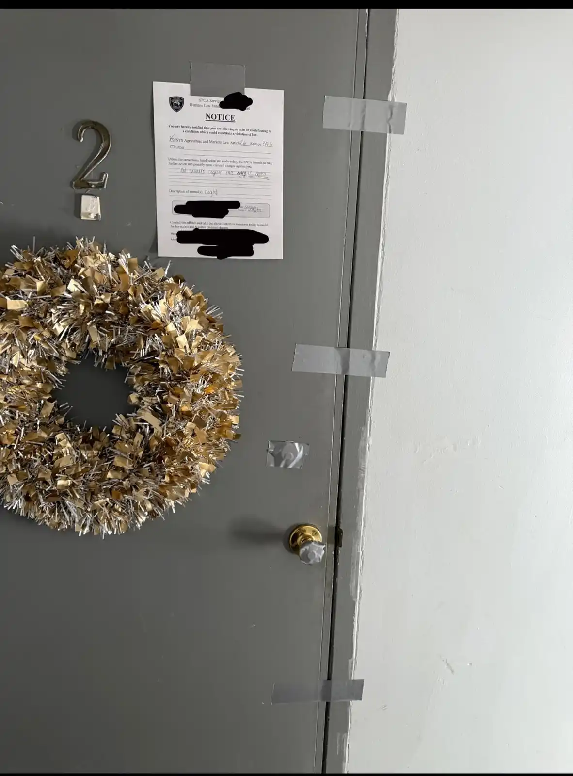 A close-up photo of a gray apartment door with a gold and silver tinsel wreath and a metal number '2' on it. Taped to the…