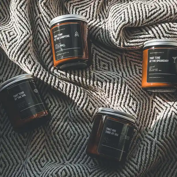 Four amber glass candles with black labels sit on a patterned woven blanket. Each candle has a nostalgic-sounding name…