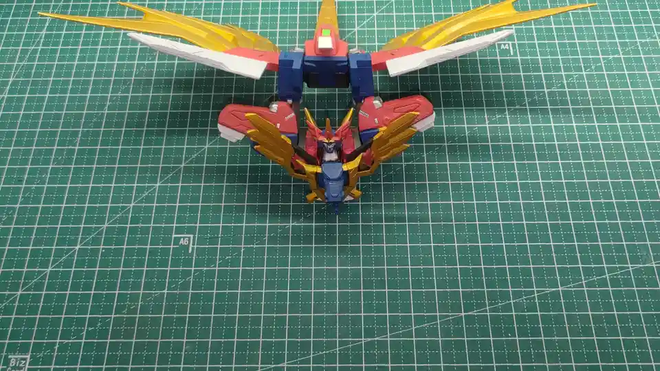 A red, blue, and gold plastic robot model with large yellow and white wings is shown from above on a green grid cutting mat…