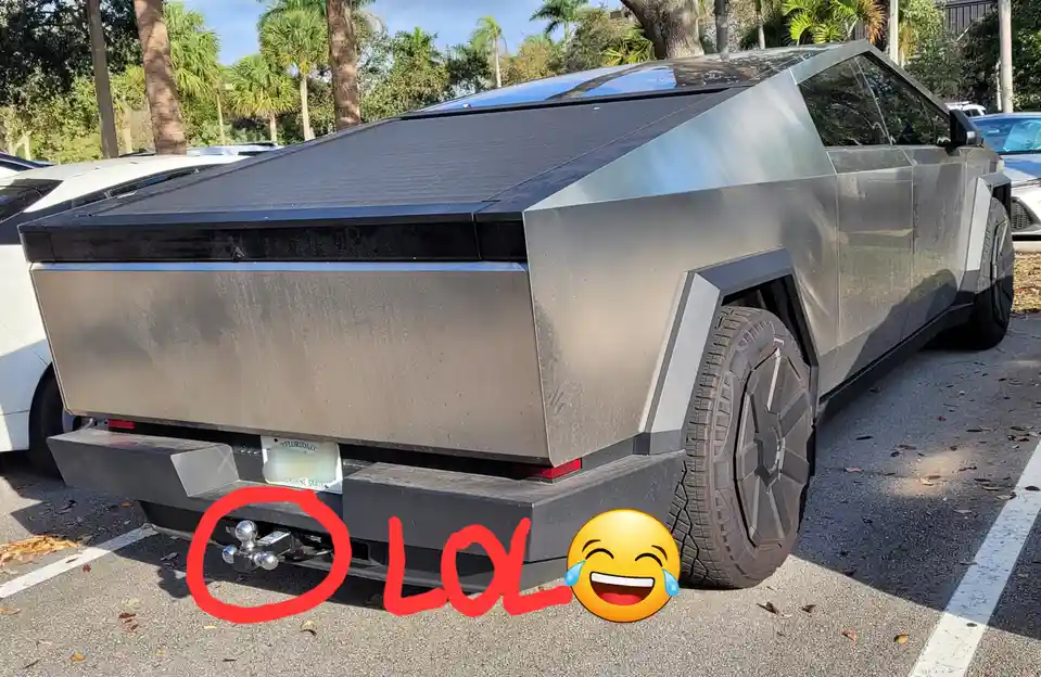 A silver Tesla Cybertruck is parked in a lot with palm trees in the background, its rear bumper has a hitch ball installed…