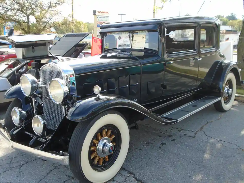 A glossy black 1931 Cadillac LaSalle sedan with white-walled tires and chrome accents is parked outdoors at a car show, its…
