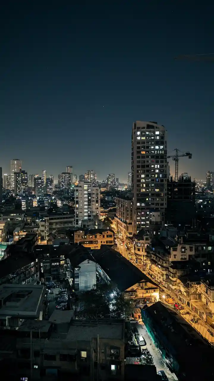 A nighttime cityscape photo showing a mix of modern high-rises and older, densely packed buildings with lights on in many…