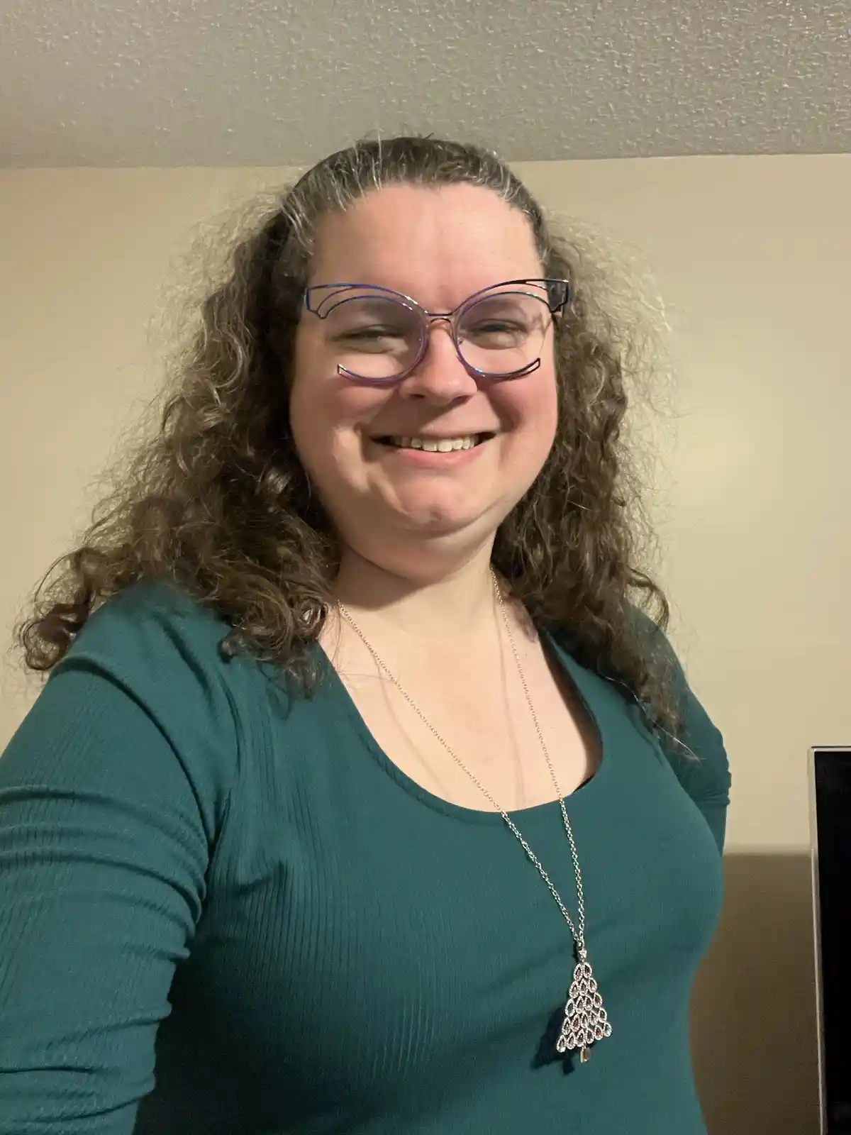 A smiling woman with curly brown hair and purple-framed glasses wears a teal ribbed long-sleeve shirt and a long silver…