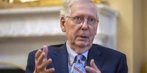 A close-up photo of an older white man with glasses, wearing a dark suit and tie, gesturing with both hands as if speaking…