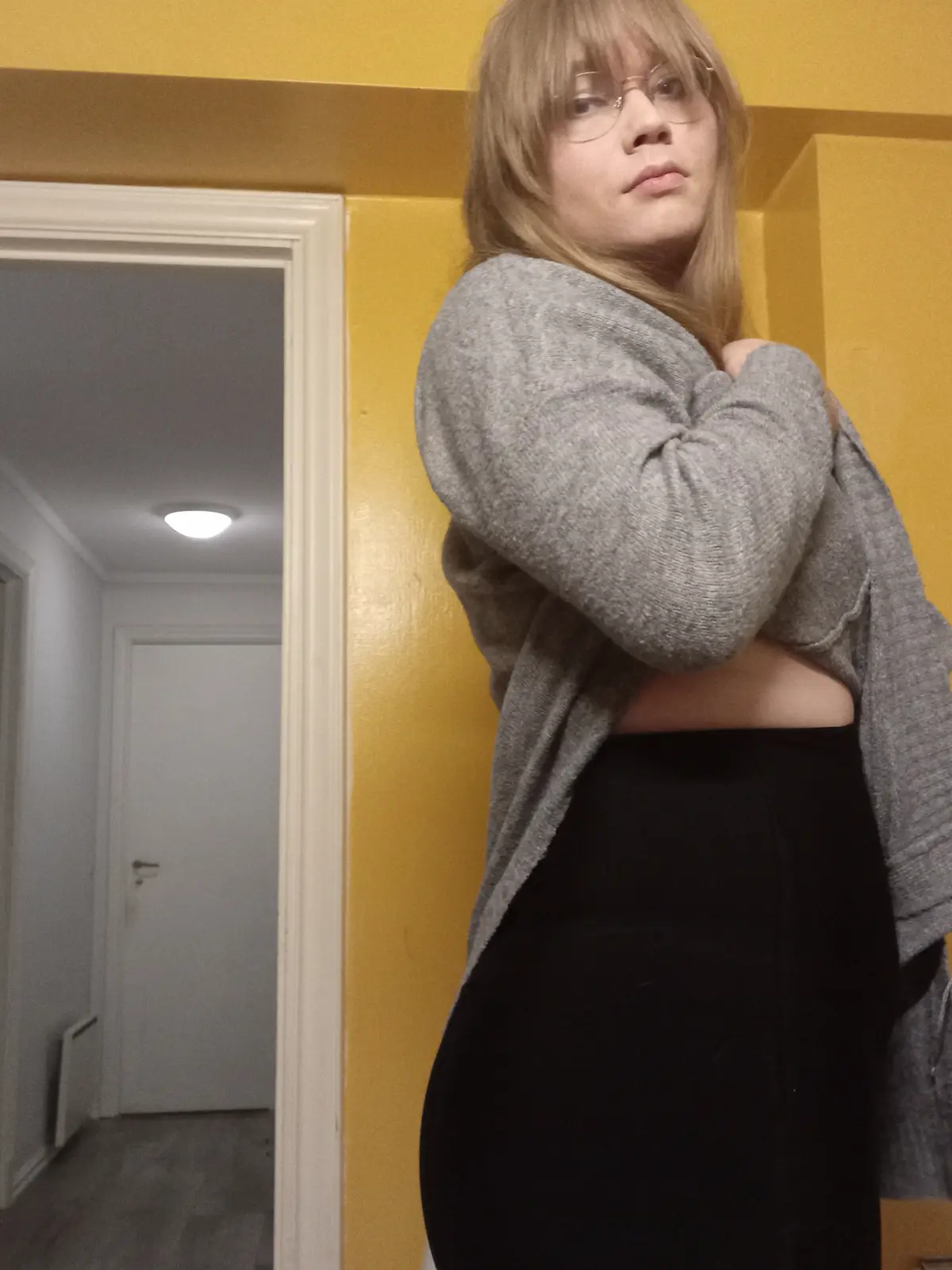 A person with blonde hair and glasses stands indoors against a yellow wall, wearing a grey sweater and black pants, arms…