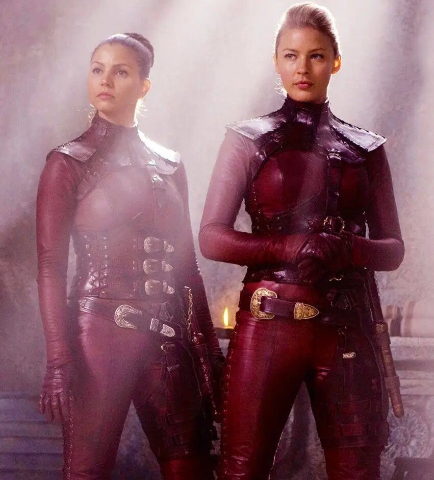 Two women in matching dark red leather outfits stand side by side in a misty, dimly lit setting with a candle behind them…