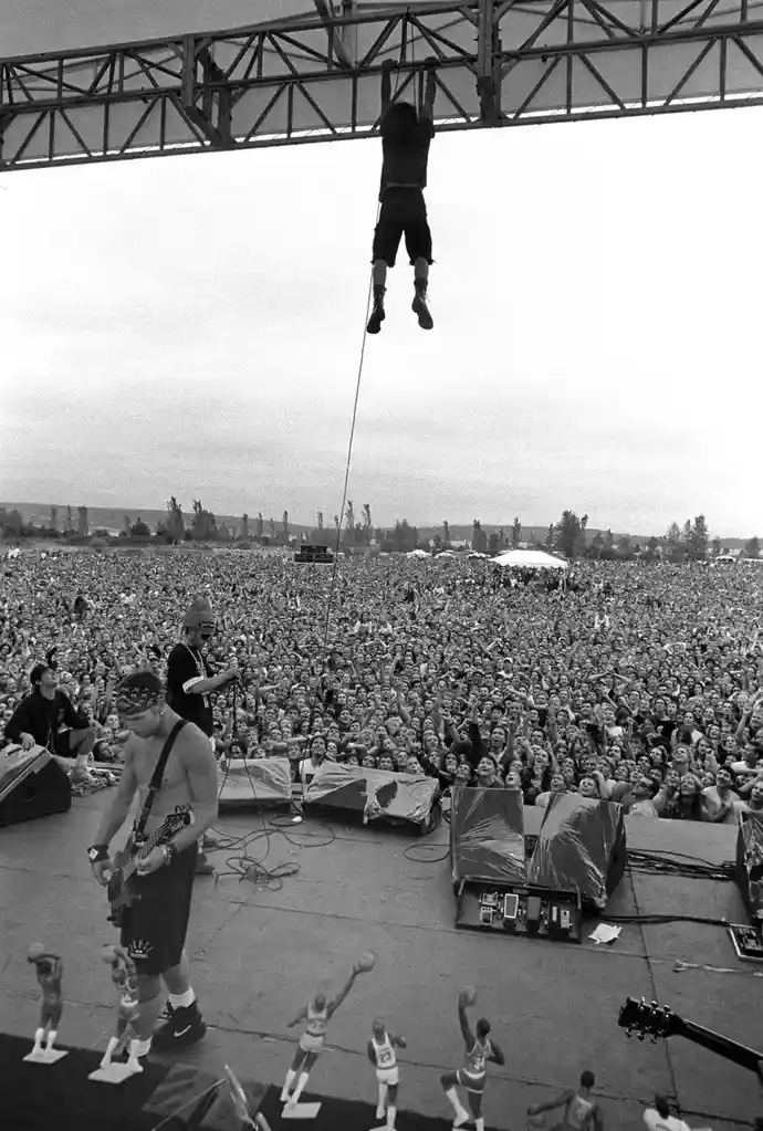 A black-and-white photo captures a live concert with a massive crowd. On stage, a shirtless guitarist plays while a man…