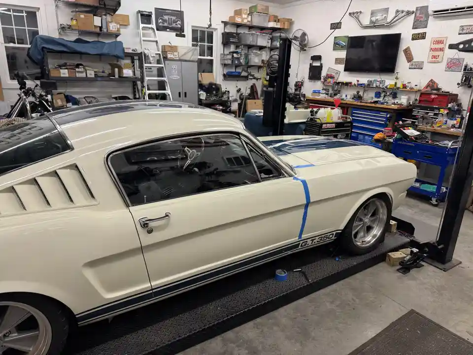 A cream-colored classic Ford Mustang with blue racing stripes and 'G.T.350' lettering is parked on a lift in a cluttered…