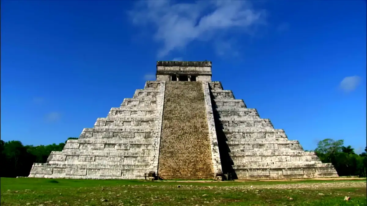 A wide-angle photograph shows a large, ancient stepped pyramid made of stone blocks under a bright blue sky with scattered…