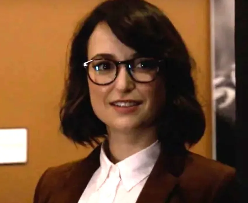 a woman with dark hair and glasses, wearing a white collared shirt and brown blazer, smiles slightly while looking forward…