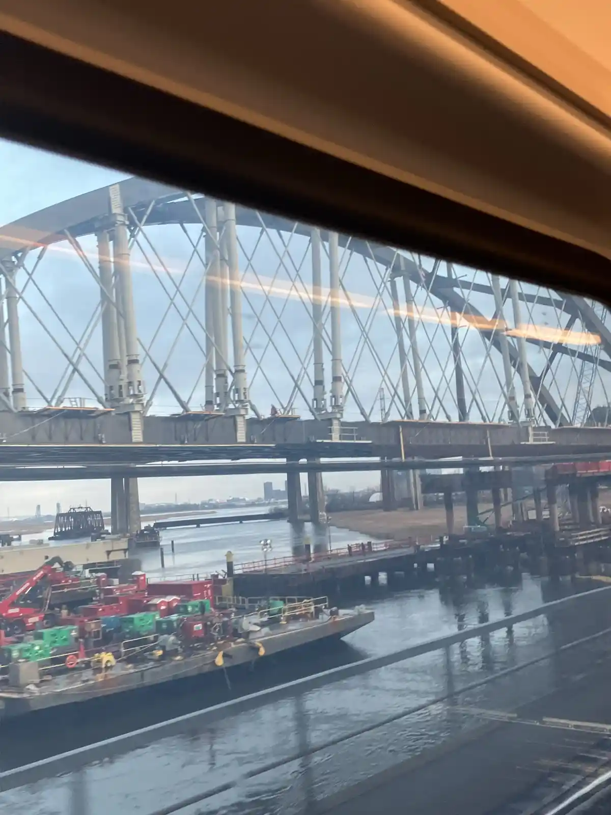 A photo taken from inside a moving vehicle, looking out a window at a large steel truss bridge under construction over a…