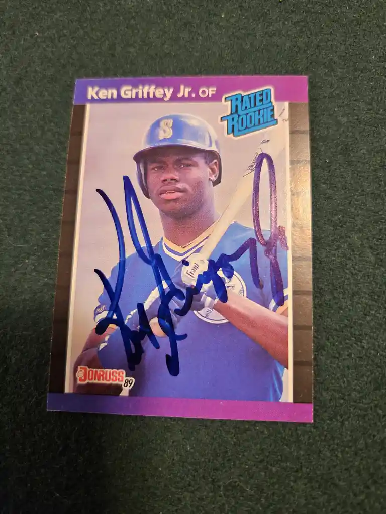 A 1989 Donruss baseball trading card of Ken Griffey Jr., labeled 'Rated Rookie', is shown with a large blue autograph across…