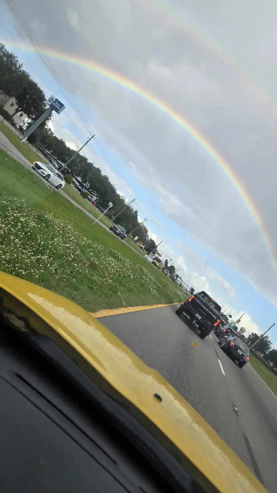 A fisheye-lens photo taken from inside a yellow car shows a bright, full rainbow arcing across a cloudy sky above a busy…