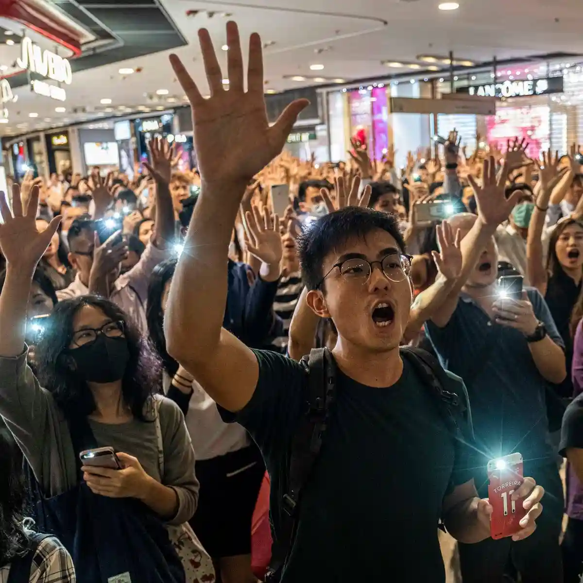 A crowd of people in a brightly lit shopping mall, many holding up phones with flashlights on, raising their hands in…