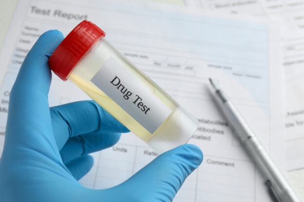 A hand wearing a blue nitrile glove holds a clear plastic vial with a red cap, labeled 'Drug Test', containing a yellowish…