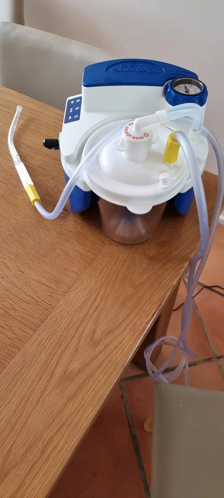 A medical-grade vacuum pump device, branded 'Vacu-Aide esu', sits on a wooden table with tubing and a clear cup underneath…