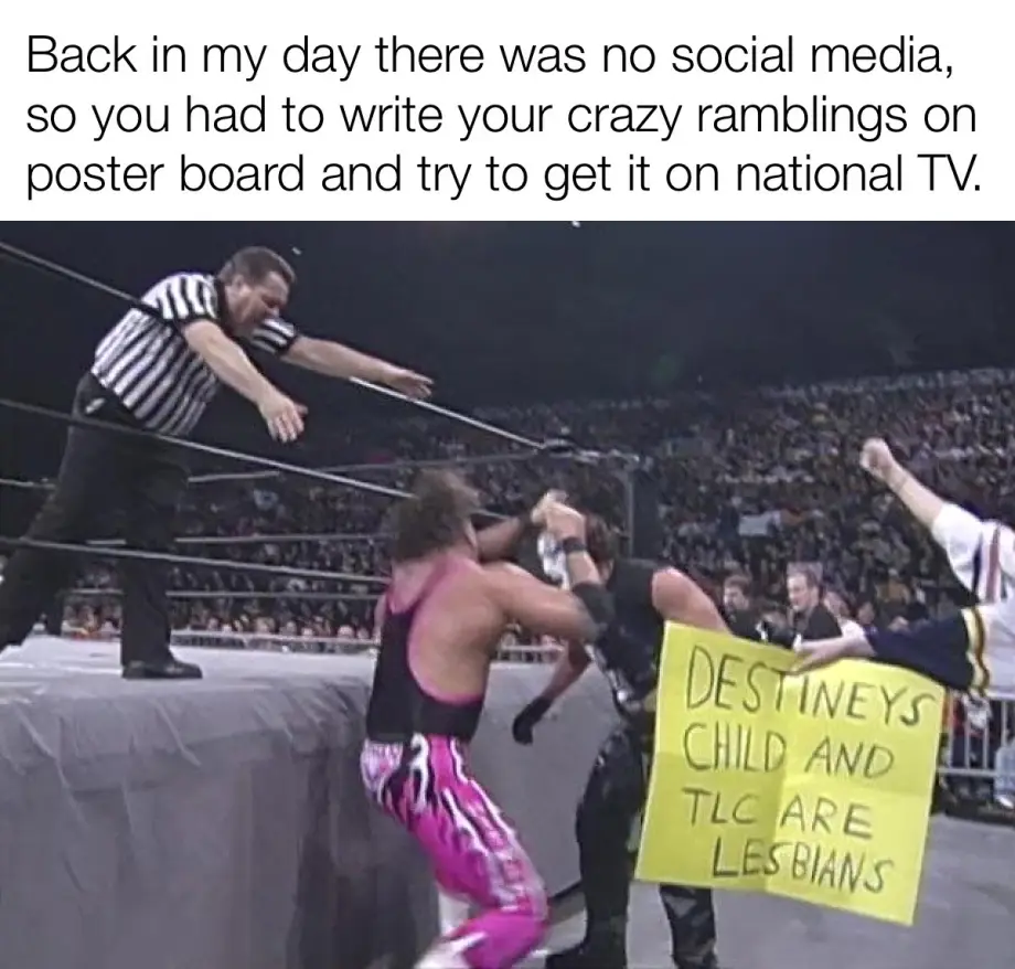 A wrestling referee leans over the ring ropes as two wrestlers grapple, while a fan holds up a yellow poster board with…