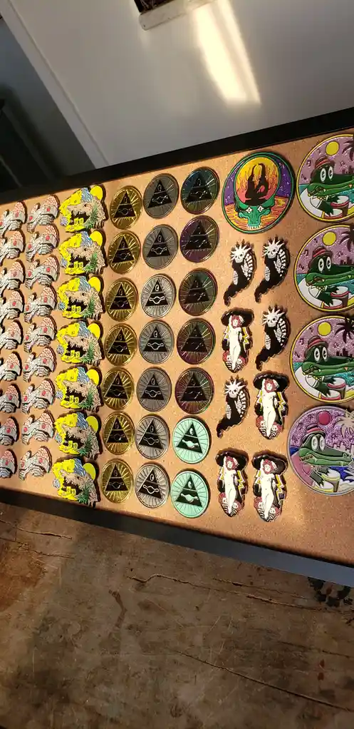 A corkboard displays a collection of enamel pins, including multiple identical pyramid-with-eye designs in various metallic…
