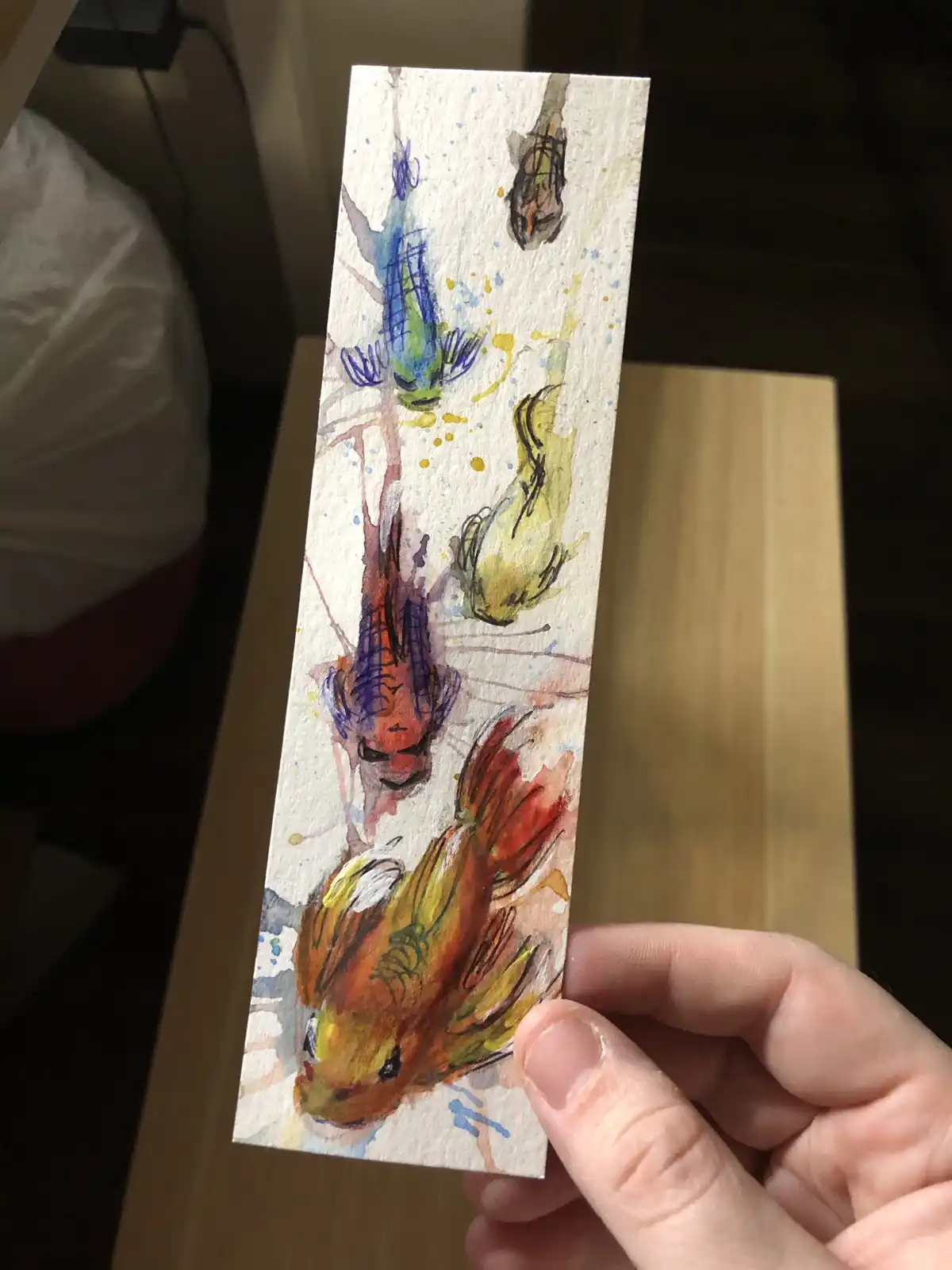 A hand holds up a small, vertically oriented watercolor painting on textured paper depicting several stylized, colorful…