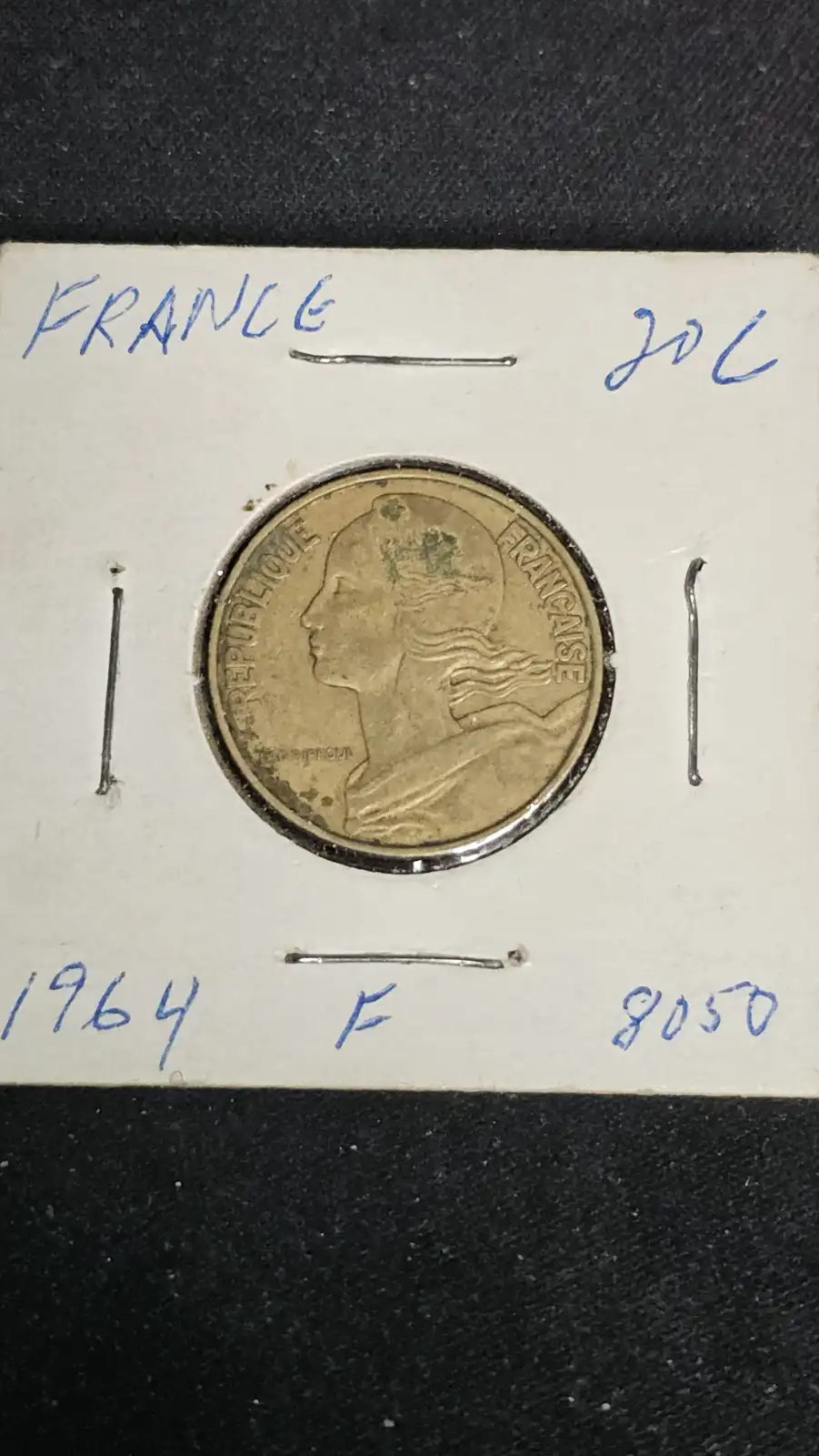 A close-up of a French 20-centime coin from 1964, housed in a white cardboard holder with handwritten details. The coin…