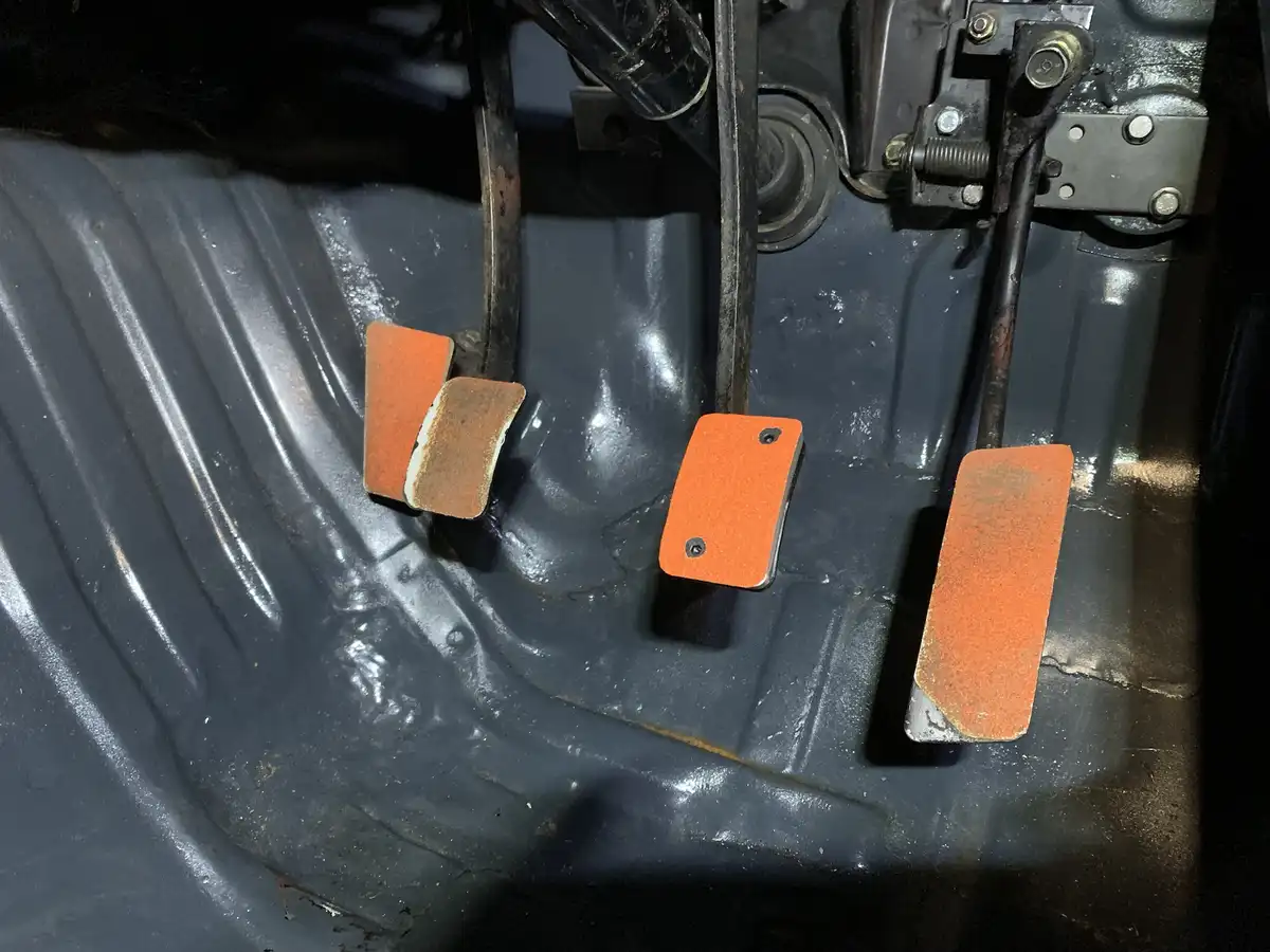 A close-up photo shows three orange pedals inside a vehicle's footwell: a worn clutch pedal on the left, a central brake…
