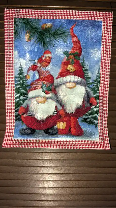 A diamond painting depicts two figures, one resembling Santa Claus and the other a gnome, both wearing red hats and red…