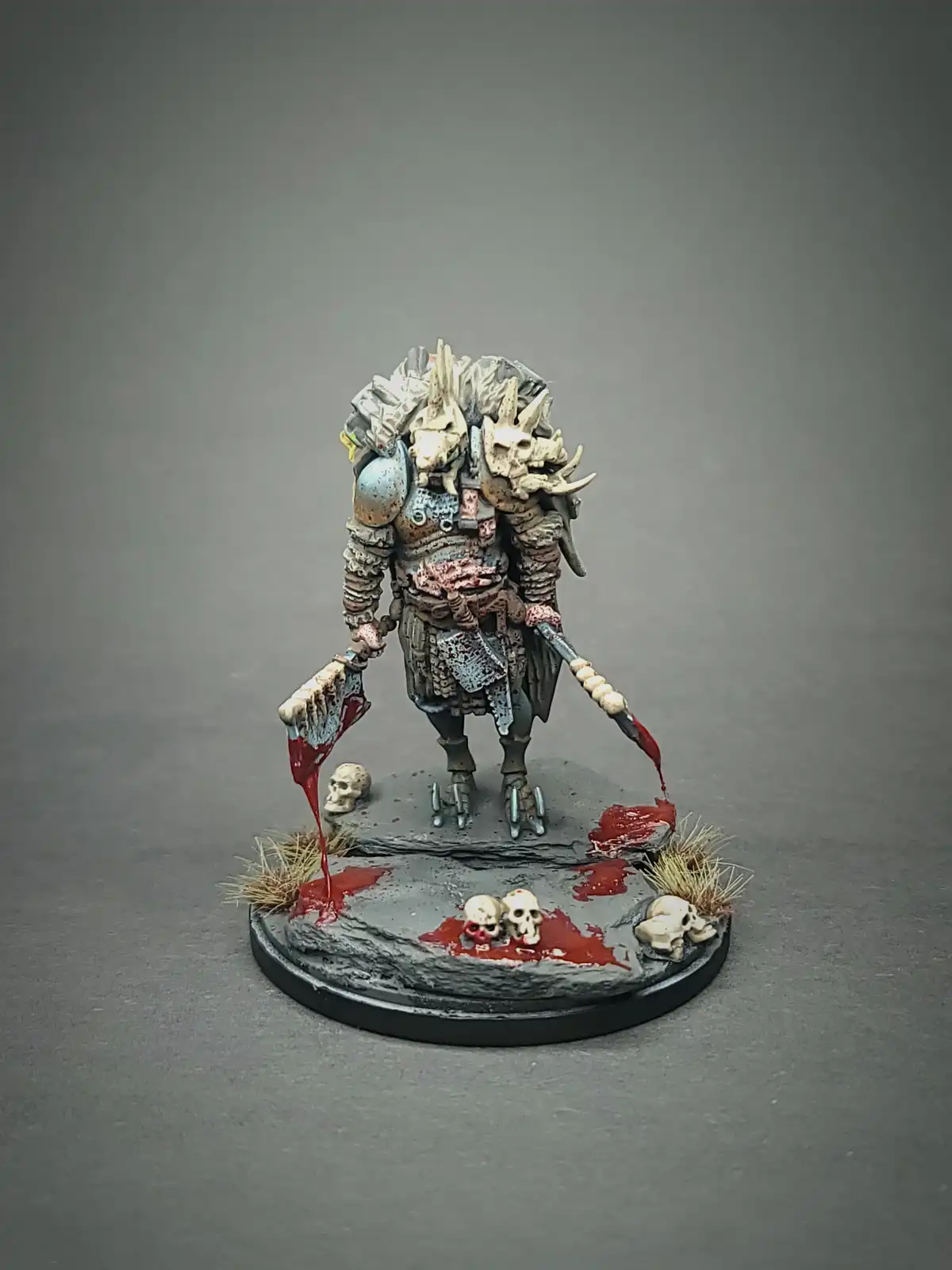 A heavily armored, grotesque fantasy warrior miniature stands on a rocky base, wielding two bloodied axes dripping red…