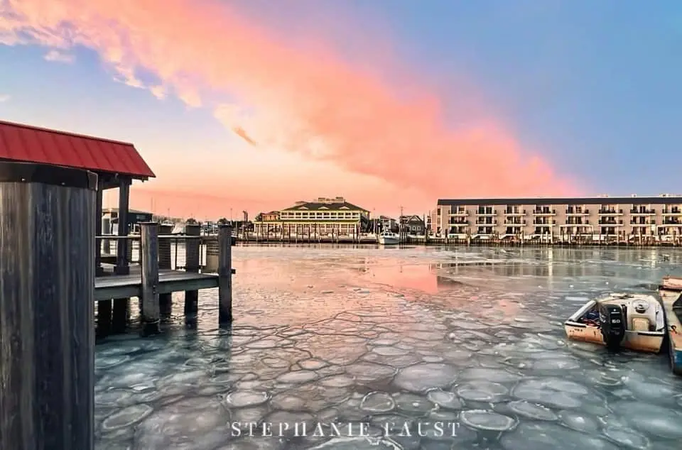 A scenic photo of a harbor at sunset with pink and blue skies, showing ice floes on the water, a wooden pier on the left…