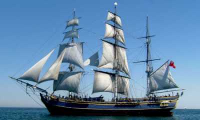 A large, multi-masted sailing ship with white sails and a red flag flies on calm blue water under a clear sky. The ship is…