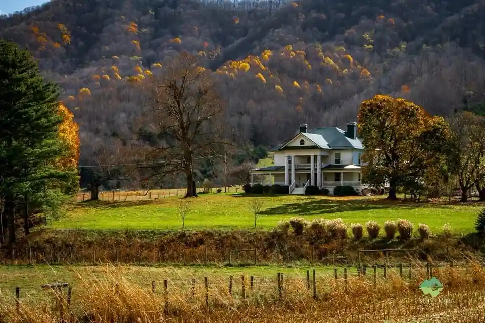 A picturesque rural house with a green roof and white columns sits on a grassy lawn, surrounded by autumn trees with orange…