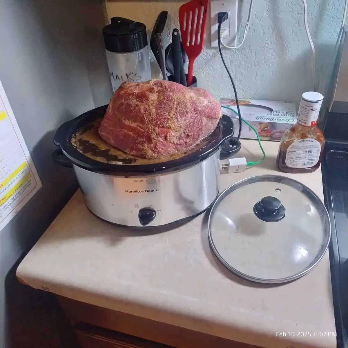 A large, raw, spice-rubbed piece of meat sits on top of an unplugged Hamilton Beach slow cooker, which is itself sitting on…
