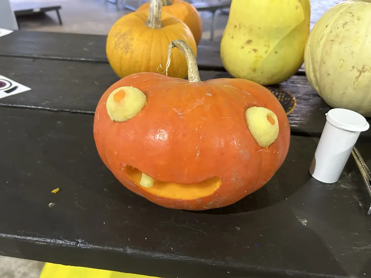 A small orange pumpkin with a carved smiling mouth and two round, pale yellow eyes made from pumpkin flesh sits on a dark…