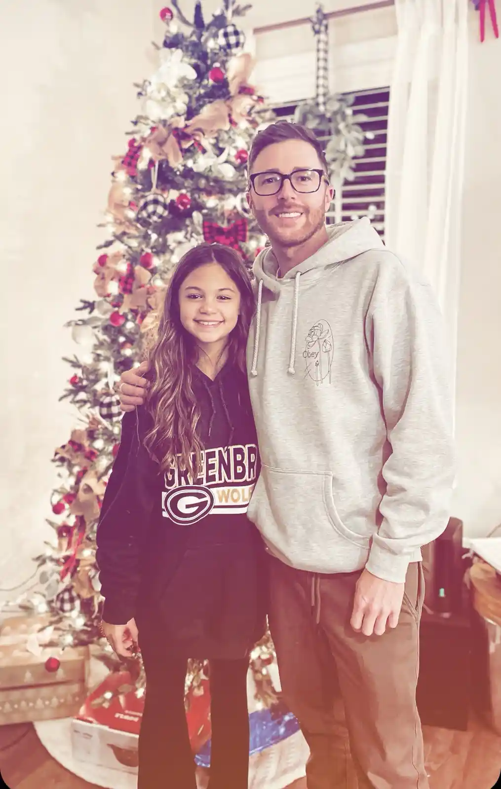 A man and a young girl stand smiling in front of a decorated Christmas tree with presents underneath. The girl wears a black…