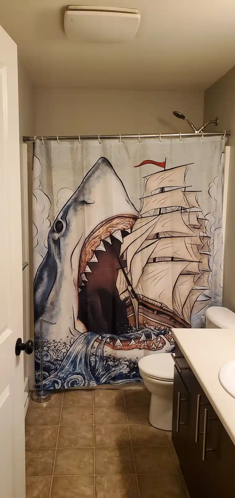 A bathroom with a shower curtain depicting a giant shark attacking a sailing ship, with a toilet and sink visible. The…
