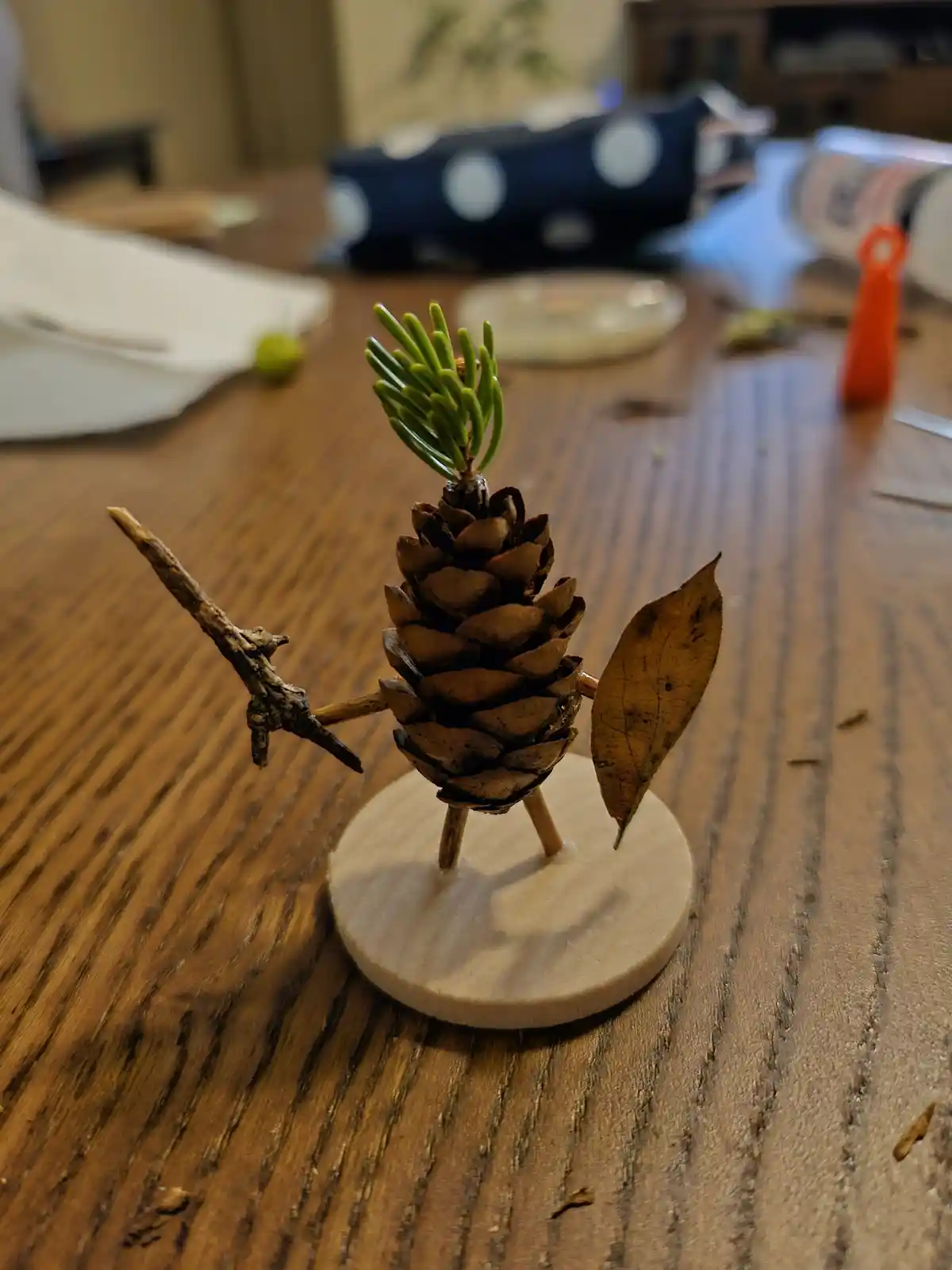 A handmade figurine crafted from a pinecone, with small twigs for arms and legs, a sprig of green pine needles for hair, and…