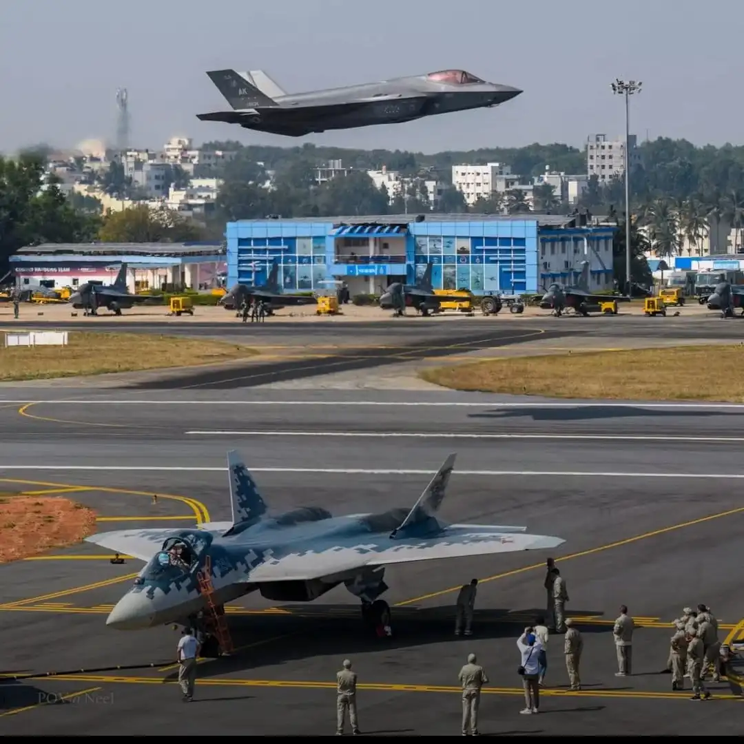 A gray F-35 fighter jet is captured mid-flight, hovering low over an airfield. Below it, a Russian Su-57 fighter jet with a…