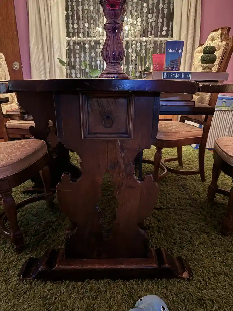 A close-up photo of an ornate, dark wooden table with a carved, asymmetrical base standing on a green shag carpet. The table…