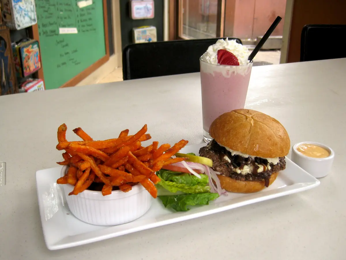 A burger with toppings, sweet potato fries, and a strawberry milkshake are arranged on a white plate and table. The burger…