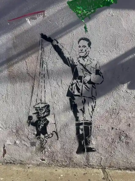 A black stencil graffiti on a textured white wall shows a man in a military-style uniform holding strings that control a…