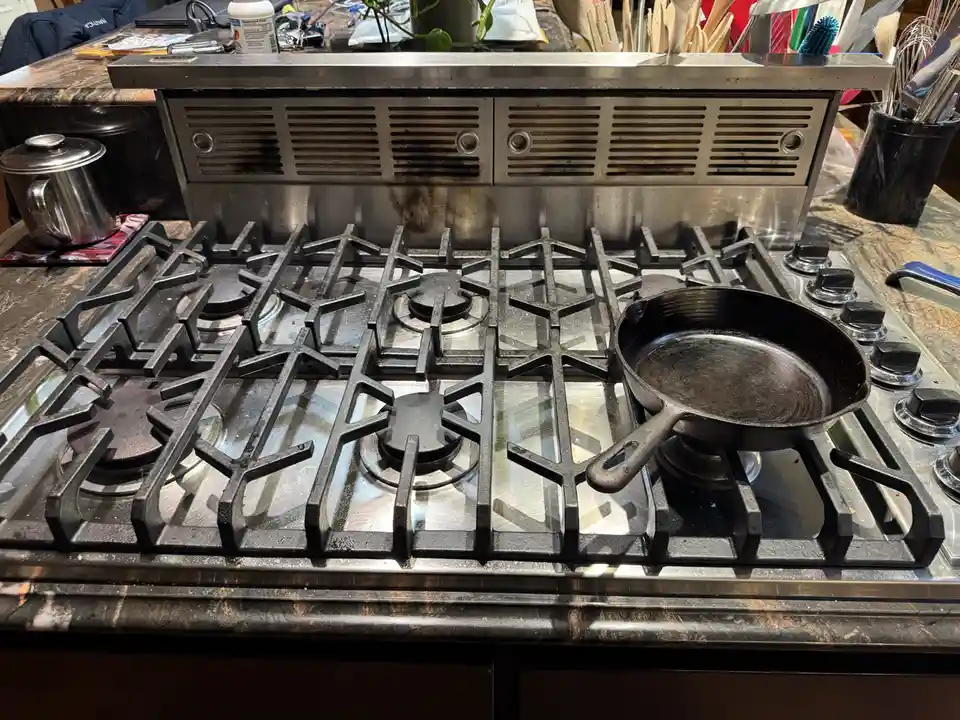 A stainless steel gas stove with black grates has a single black cast iron skillet sitting on one burner. The stove and…
