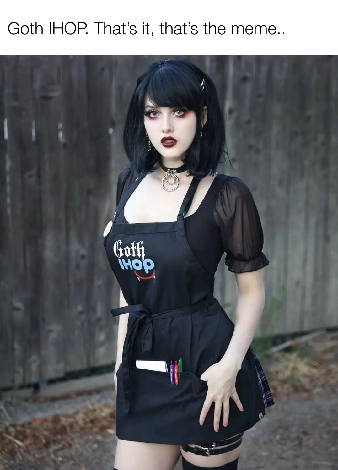 A woman with black hair, goth makeup, and a choker wears a black apron with 'Goth IHOP' printed on it, standing in front of…