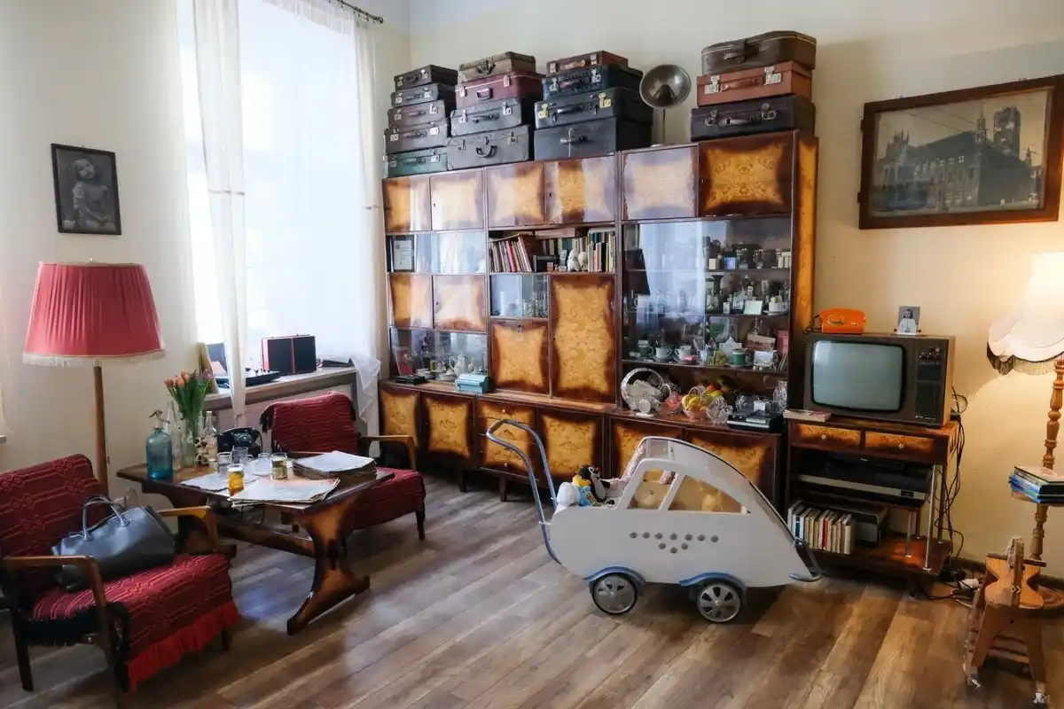 A cozy, cluttered living room with vintage furniture, including a large wooden cabinet topped with stacks of old suitcases…